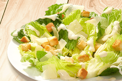 Caesar-Salat auf weißem Teller: Römersalatherzen, knusprige Croutons, Parmesanspäne und cremiges Dressing auf hellem Holzuntergrund.