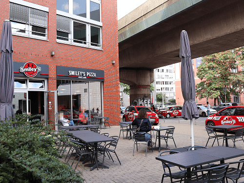 Außenansicht eines Smiley's Restaurants mit Terrasse, Sitzplätzen und parkenden Lieferfahrzeugen