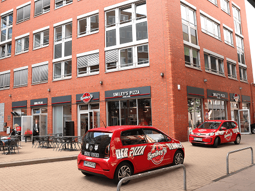 Eckgebäude mit Smiley's Filiale, roten Lieferautos und Sitzmöglichkeiten vor dem Eingang