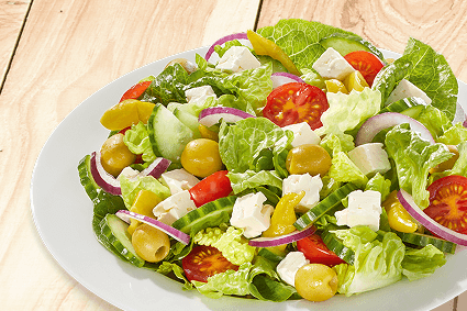 Griechischer Hirtensalat auf weißem Teller: Römersalat, Gurkenscheiben, Cherrytomaten, grüne Oliven, rote Zwiebelringe, Fetawürfel und Peperoni auf hellem Holzuntergrund.