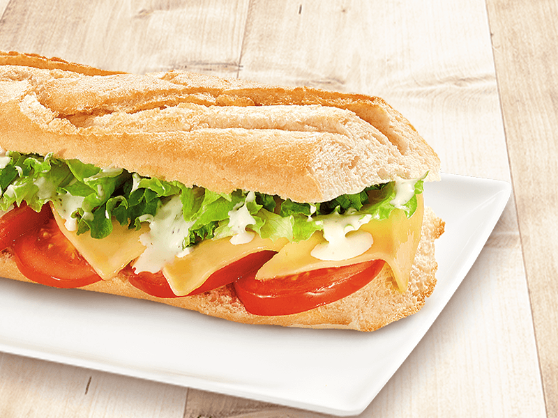 Belegtes Baguette mit Käse, Tomaten, Salat und Sauce auf weißem Teller vor hellem Holzhintergrund.