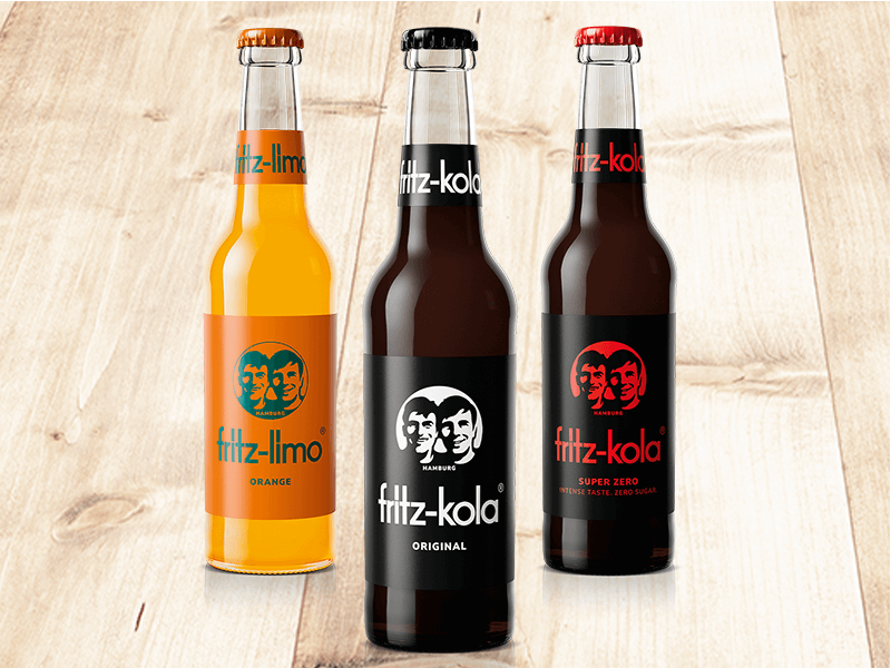 Drei fritz-Getränkeflaschen: fritz-limo Orange, fritz-kola Original und fritz-kola Super Zero auf hellem Holzhintergrund.