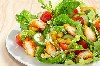 Frischer Salat mit knusprigen Hähnchenstreifen, Rucola, Tomaten, Gurken und Mais auf einem weißen Teller. Hintergrund in heller Holzoptik.