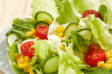 Gemischter Salat auf einem weißen Teller: Römersalat, Blattsalate, Cherrytomaten, Gurkenscheiben und Mais auf hellem Holzuntergrund