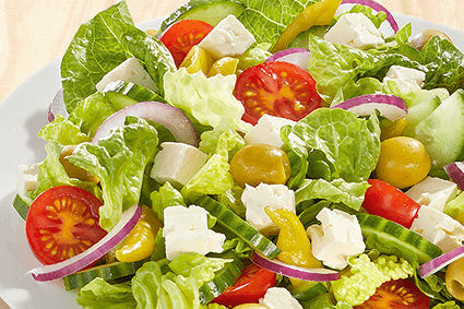 Griechischer Hirtensalat auf weißem Teller: Römersalat, Gurkenscheiben, Cherrytomaten, grüne Oliven, rote Zwiebelringe, Fetawürfel und Peperoni auf hellem Holzuntergrund.