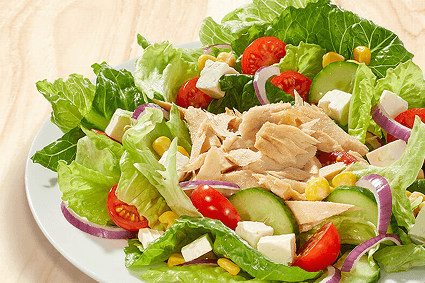 Thunfischsalat auf weißem Teller: Römersalat, zarte Thunfischstücke, Gurkenscheiben, Cherrytomaten, Fetawürfel, Mais und rote Zwiebelringe auf hellem Holzuntergrund.